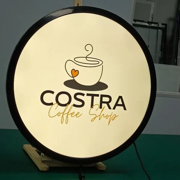Biển vẫy hộp đèn hình tròn nổi bật với thiết kế đèn sáng, logo ly cà phê và chữ “COSTRA Coffee Shop” rõ nét, tạo điểm nhấn thu hút cho quán